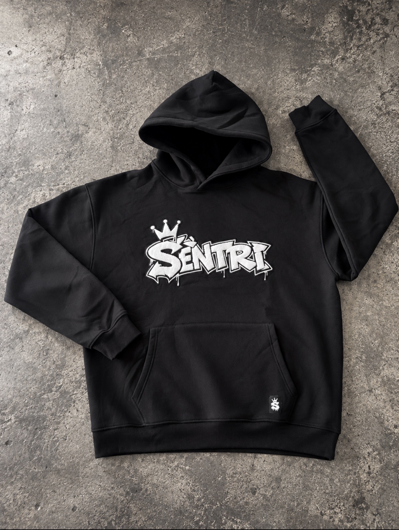 SÈNTRI Drop 2 black hoodie laid flat on concrete